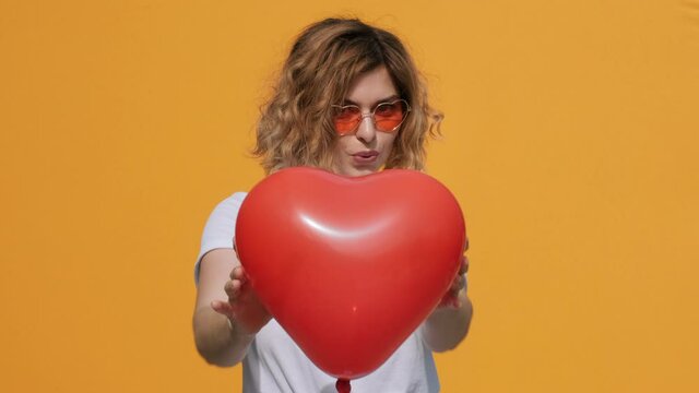Portrait of happy woman with red heart balloon holding in her hands imitating how heart beats in front of herself smiling at camera on yellow background summer. Emotions Positive girl Flirt. Valentine