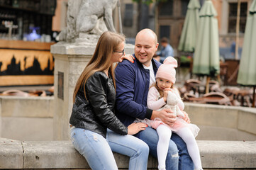 Parents and their little daughter are sitting at the old city fountain.