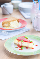 Two pieces of cakes on a colorful plates and wooden table. Cheesecake with red jelly top and mint. Blurred background. Cozy restaurant scene