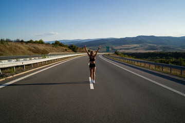 Young girl running along the road. Athletic happy woman jogging in trendy black sexy top and shorts enjoying the sun exercising. Healthy lifestyle. Perfect fitness body shapes and tan skin