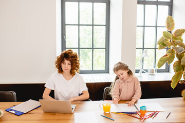 Obraz na płótnie Canvas redhead freelancer mother using laptop near glass of orange juice and cute daughter drawing at home