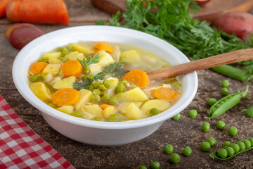 Spring vegetable soup with peas, young potatoes and carrots served in white bowl. Healthy vegan soup with ingrediants on rustic wooden table.