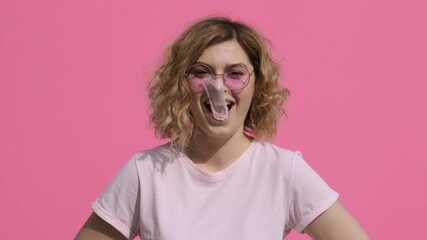 Portrait of woman happy smile blows up bubble from chewing gum and rips it slow motion pink t-shirt, pink sunglasses, curly hair on pink background summer. Monotone. Emotions. Lifestyle. Positive girl