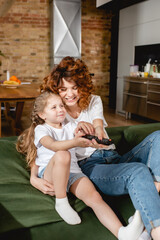 cute kid holding remote controller and sitting near cheerful mother on sofa