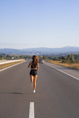Obraz premium Young girl running along the road. Athletic happy woman jogging in trendy black sexy top and shorts enjoying the sun exercising. Healthy lifestyle. Perfect fitness body shapes and tan skin