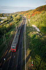 Fototapeta na wymiar View from the top of the railway. Passenger train rides on the railway tracks. Colorful forest along the road. Gold autumn.