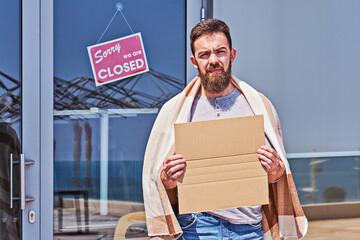 Unemployed guy with empty signboad near closed door office or cafe. Unemployment during coronavirus...