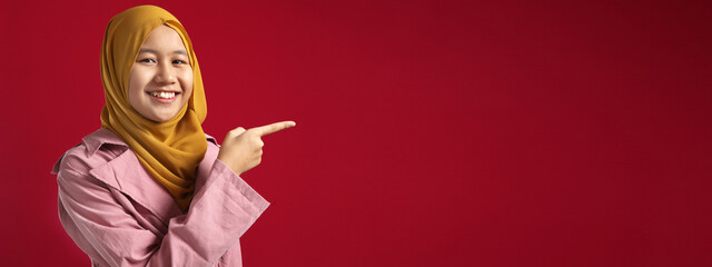 Cheerful young Asian muslim teenage girl wearing hijab looking to cmera and pointing on side on the red background, horizontal banner with copy space