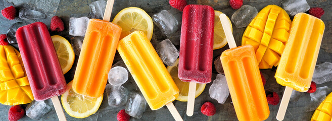 Variety of colorful summer popsicles with ice and fruit. Top view close up on a dark slate banner background.