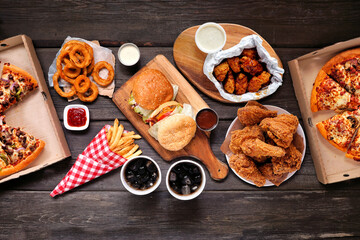 Fototapeta Table scene with large variety of take out and fast foods. Hamburgers, pizza, fried chicken and sides. Above view on a dark wood background. obraz