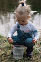 Little girl in rubber boots catches and feeds fish on the river in a jar