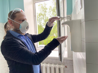 A man in a respirator and medical gloves treats his hands with an antiseptic. A man clicks on a dispenser for an antiseptic in a hospital after receiving a doctor.