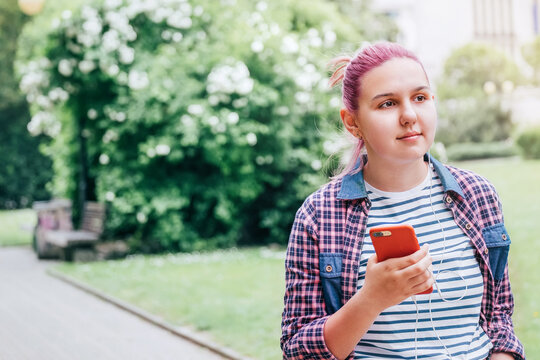 Portrait of Beautiful modern smiling young female teenager in a checkered shirt listening music using smartphone and earphones as she walking in park. Modern teens in digital world concept image.