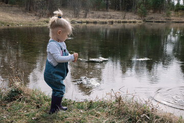 Fototapeta na wymiar Little girl in rubber boots catches and feeds fish on the river in a jar