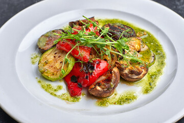 Grilled baked vegetables, eggplant, zucchini, bell pepper, champignon mushrooms, on a white plate against a dark background, with microgreen