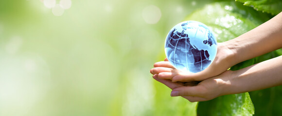 World Environment Day or Earth Day concept. Blue glass globe in woman hands on blurred green leaves banner background. Saving planet, save, protect clean nature and ecology, sustainable lifestyle.