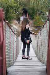 Beautiful woman with brunette hair in dark clothes and sunglasses. Fashion street photography. Fashion model is standing on suspension bridge on nature background.