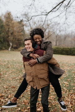 A white man giving a black woman a piggy back on a leaf strewn lawn and laughing.