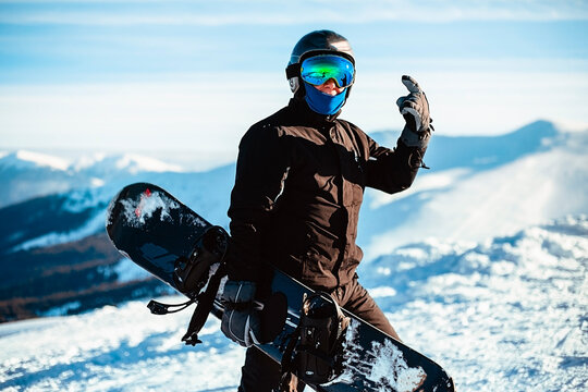A person wearing a black ski suit, helmet and goggles holding a snowboard standing on top of a snowy mountain.