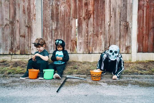 Three children dressed for Halloween, one as a skeleton, sitting at the side of a road with their buckets of sweets.