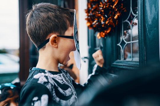 Close Up Of A Boy Wearing A Mask Knocking On A Front Door At Halloween.