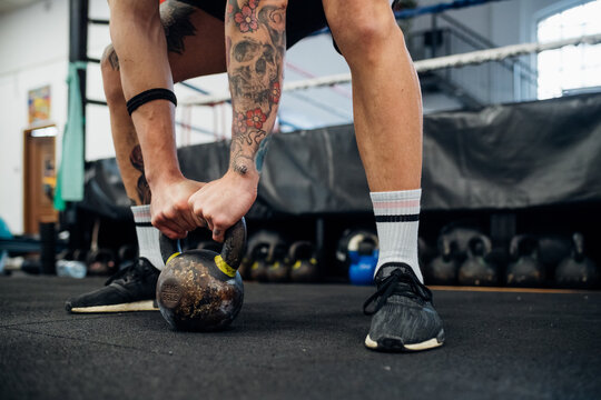Tattooed arms and legs of a man holding a kettle bell on the floor.