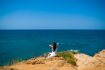 A romantic young girl brunette in a long white dress sits on the edge of the island 's land. Beautiful landscape of exotic white sand sea beach Southeast Asia, Thailand. View from the mountain.