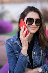 Attractive girl resting on nature at weekend. Girl in jeans jacket sitting in park. Caucasian brunette young woman talking on smartphone outside alone.Technologies and gadgets.