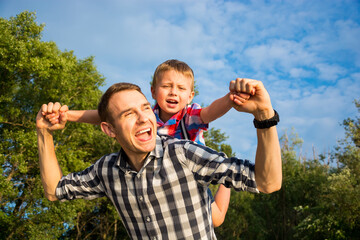 Happy young father holds his son piggyback ride on his shoulders and looking up. Little boy is...