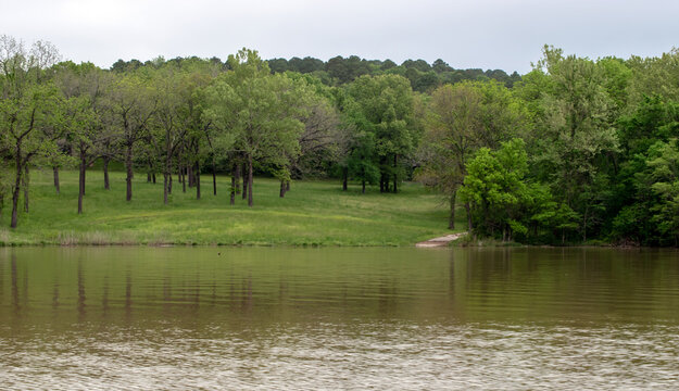A small walkway is seen at the edge of the waters of tranquil and peaceful lake Eucha in Oklahoma with a grove a green trees and still waters.
