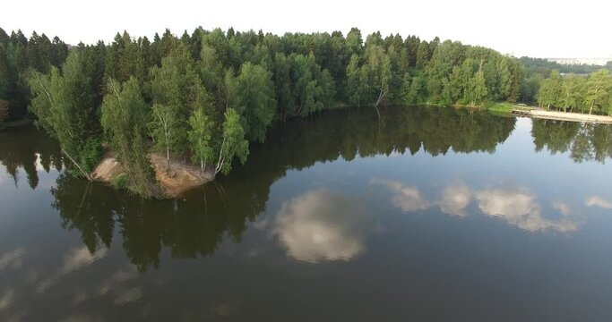 4K aerial summer early morning high quality video footage of pristine lush green pine tree surrounded forest lake with dark calm water reflecting clouds, a dam, island and a sand beach near small town