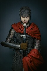 Gorgeous young woman in knight armour and steel chainmaille holding sword on dark background....