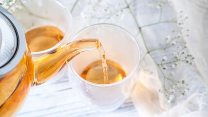 From transparent glass teapot pour golden tea in glass mug. Glass teapot pouring black tea into cup. White wooden table