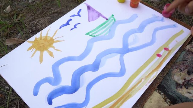 Closeup top view video footage of preschool boy of 5 years old having fun outdoor sitting on picnic blanket on ground. Child painting cute picture of sunny summer beach with sailing boat in blue sea.