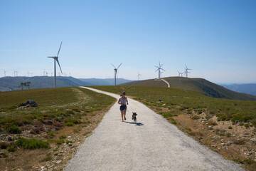 Fototapeta premium Woman girl running with dog on road landscape at Serra da Freita Arouca Geopark wind turbines landscape, in Portugal