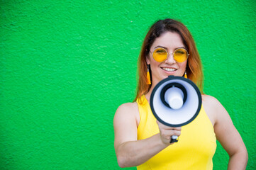 Fototapeta premium Happy woman in a yellow dress glasses and earrings on a green background shouting into a megaphone. Portrait of a girl holding a loudspeaker. Lady with a perfect snow-white smile.