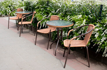 Summer street cafe. The glass table and brown chairs are outdoors. A small cozy restaurant with retro furniture and bushes with white flowers.