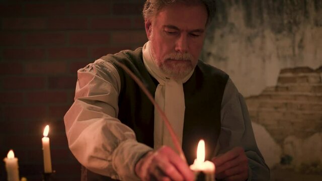 In a scene from the 18th century a man sitting at rustic desk uses a feather quill pen to write a letter in a room lit only by candlelight and other firelight.
