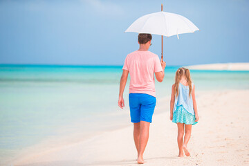 Young father and his little daughter walking under umbrella on white sand beach