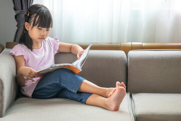 Asian little girl thinking while reading a book on the sofa in the room.