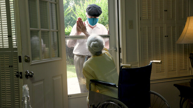 An Elderly Woman In A Wheelchair Social Distancing Because Of COVID19 Visits With A Caring Relative Or Neighbor Through Her Glass Storm Door.