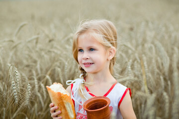 Portrait of a little girl with a brown jug and bread in her hand. A girl poses in a wheat field in...