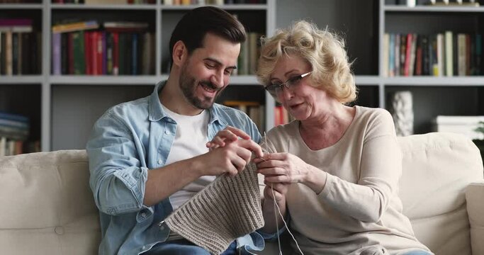 Happy middle aged mother teaching helping grownup son knitting warm winter clothes. Smiling young man learning creating handmade woolen scarf or sweater, enjoying hobby time with older mother at home.