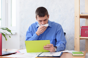 A man in the office with a tablet in his hand coughs in a napkin