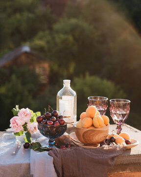 Summer still life in the village. In a blue vase lies cherry berries, apricots in a wooden plate, pink roses in a vase. Empty bottle of cambuca. Two purple glasses. Relaxing in nature. Sun rays.