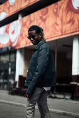 Fototapeta premium Young millennial african boy in city. Happy black man in jacket with camera. Generation Z concept. Man walking on the street.Portrait of pensive modern afro man with sunglasses and camera