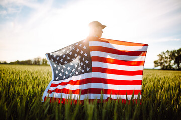 Young patriotic farmer stands among new harvest. Boy walking with the american flag on the green wheat field celebrating national independence day. 4th of July concept. - obrazy, fototapety, plakaty