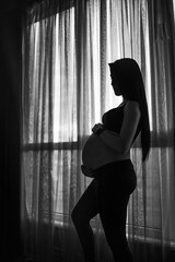 Pregnant woman in black clothes holds hands on belly on a dark background. Pregnancy, maternity, preparation and expectation concept - close up of happy pregnant woman with big belly at window.