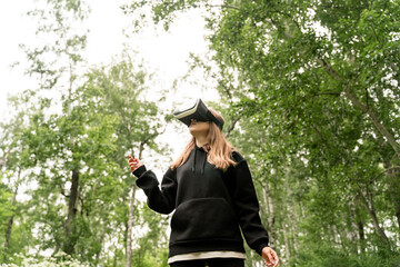 girl stands in the middle of the forest in virtual glasses is surprised stretches her hand to the top