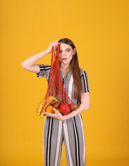 Portrait of a beautiful young woman with long hair holding a net with fruits in her hands. Standing straight, wearing a striped jumpsuit. On an orange background